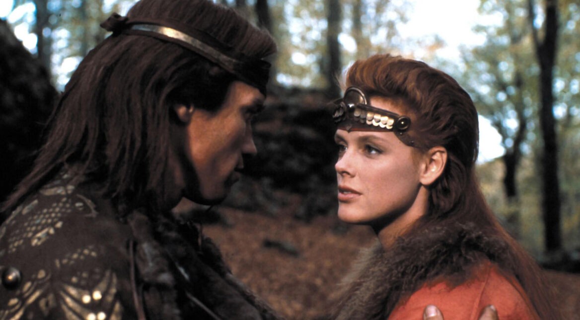 Auf dem Bild im Red Sonja Reboot Artikel siehst Du ein Bild aus dem Originalfilm „Red Sonja“ von 1985 sind Brigitte Nielsen als Red Sonja und Arnold Schwarzenegger als Kalidor in einer Waldszene zu sehen.