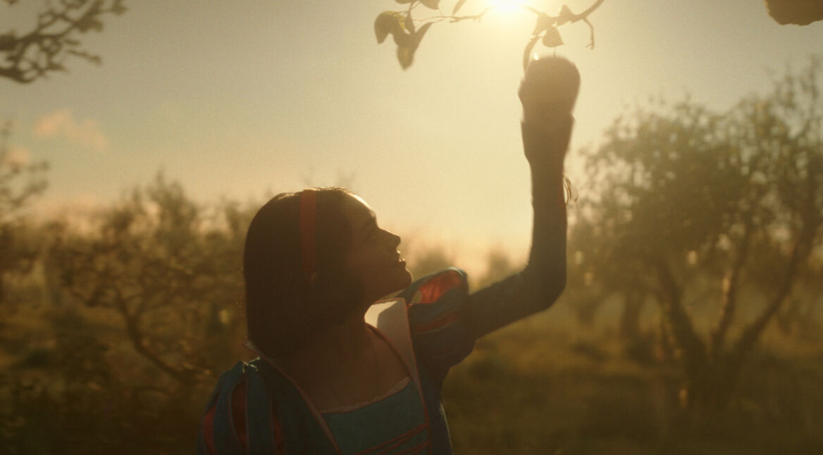 Auf dem Bild aus Schneewittchen (2025) ist Rachel Zegler als Schneewittchen in einem märchenhaften Obstgarten zu sehen. In sanftem, goldenen Licht greift sie nach einem Apfel, während die Szenerie eine verträumte, fast magische Atmosphäre ausstrahlt. Ihr blau-rotes Kleid erinnert an das klassische Design der 1937-Version, jedoch mit realistischeren Details. Im Vergleich zum handgezeichneten Originalfilm wirkt die Neuverfilmung cineastischer und natürlicher, was den Kontrast zwischen den beiden Schneewittchen-Adaptionen betont.
