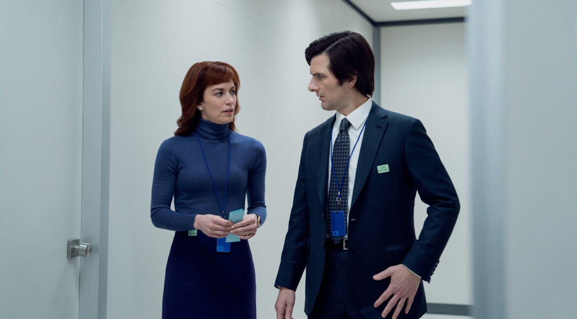 Auf dem Bild aus Severance Staffel 2 sind Adam Scott als Mark Scout und Britt Lower als Helly Riggs in einem sterilen, weiß beleuchteten Büroflur zu sehen. Mark trägt einen dunklen Anzug mit Krawatte, während Helly ein eng anliegendes blaues Rollkragenkleid trägt. Beide tragen ID-Karten an blauen Bändern und führen ein ernstes Gespräch. Severance Staffel 2 Ende erklärt – diese Szene deutet auf eine wichtige Entwicklung innerhalb von Lumon Industries hin.