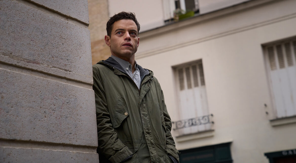 Auf dem Bild aus The Amateur ist Charles Heller zu sehen, gespielt von Rami Malek. Er steht in einer Straßenszene an einer Häuserwand und blickt mit ernstem, nachdenklichem Gesichtsausdruck in die Ferne. Sein olivgrüner Parka und die gedeckten Farben der Umgebung verleihen der Szene eine gespannte, geheimnisvolle Stimmung. Das Bild vermittelt einen stillen Moment voller innerer Anspannung und Ungewissheit.