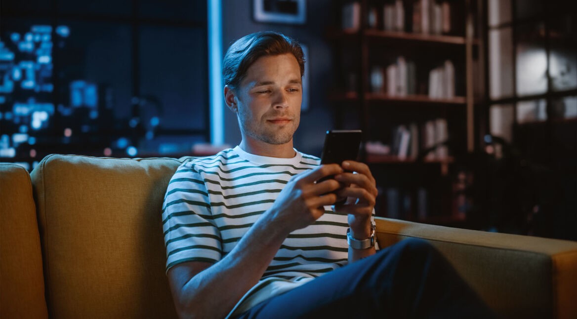 Ein Mann sitzt auf einem Sofa und hält ein Smartphone in der Hand.