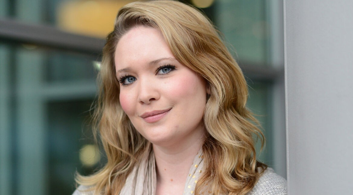 Auf dem Bild zur ACOTAR-Verfilmung ist die US-amerikanische Fantasy-Autorin Sarah J. Maas zu sehen, die „Das Reich der Sieben Höfe“ geschrieben hat. Sie blickt freundlich in die Kamera, trägt ihre blonden Haare offen und ist vor einem unscharfen Hintergrund mit Glasfassade zu sehen. Das Bild vermittelt eine ruhige, optimistische Stimmung und hebt die Autorin der Romanvorlage zur ACOTAR-Verfilmung hervor.