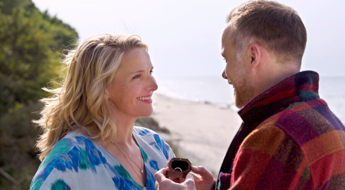 Auf dem Bild aus der ARD-Serie Praxis mit Meerblick, Teil des Programms der ARD-Serien 2025, sind Max Jensen, gespielt von Bernhard Piesk, und Nora Kaminski, dargestellt von Tanja Wedhorn, in einer romantischen Szene am Strand zu sehen. Max hält einen geöffneten Ringkasten, während beide sich verliebt ansehen. Die Stimmung ist emotional und voller Zuneigung.