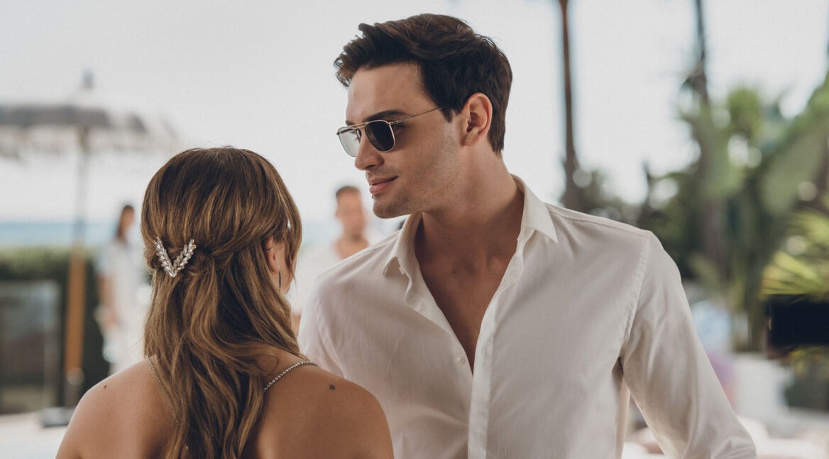 Diese Szene aus Culpa Nuestra zeigt Gabriel Guevara als Nick im weißen Hemd mit Sonnenbrille, wie er einer Frau – von hinten zu sehen, mit eleganter Haarspange – nahekommt. Die Atmosphäre wirkt sommerlich und leicht angespannt, mit Palmen und Meer im Hintergrund, was auf eine Hochzeit oder Feier am Strand hindeutet.