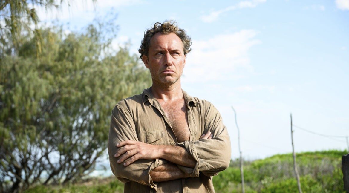 Das Bild zum Thema Eden Ende erklärt zeigt Jude Law als Dr. Friedrich Ritter in einer entschlossenen Pose mit verschränkten Armen. Sein wettergegerbtes Gesicht und das offene Hemd unterstreichen die Härte und Isolation der Figur. Im Hintergrund ist tropische Vegetation zu sehen, was die raue Umgebung ihres Eden-Projekts widerspiegelt.