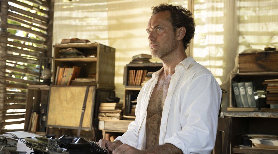 Jude Law verkörpert in Eden den Charakter Dr. Friedrich Ritter und ist hier an einer Schreibmaschine zu sehen, umgeben von Holzmöbeln und alten Büchern. Der konzentrierte Blick und die schlichte Umgebung spiegeln die isolierte und intellektuell geprägte Persönlichkeit wider, die im Zentrum der Eden wahre Geschichte steht.