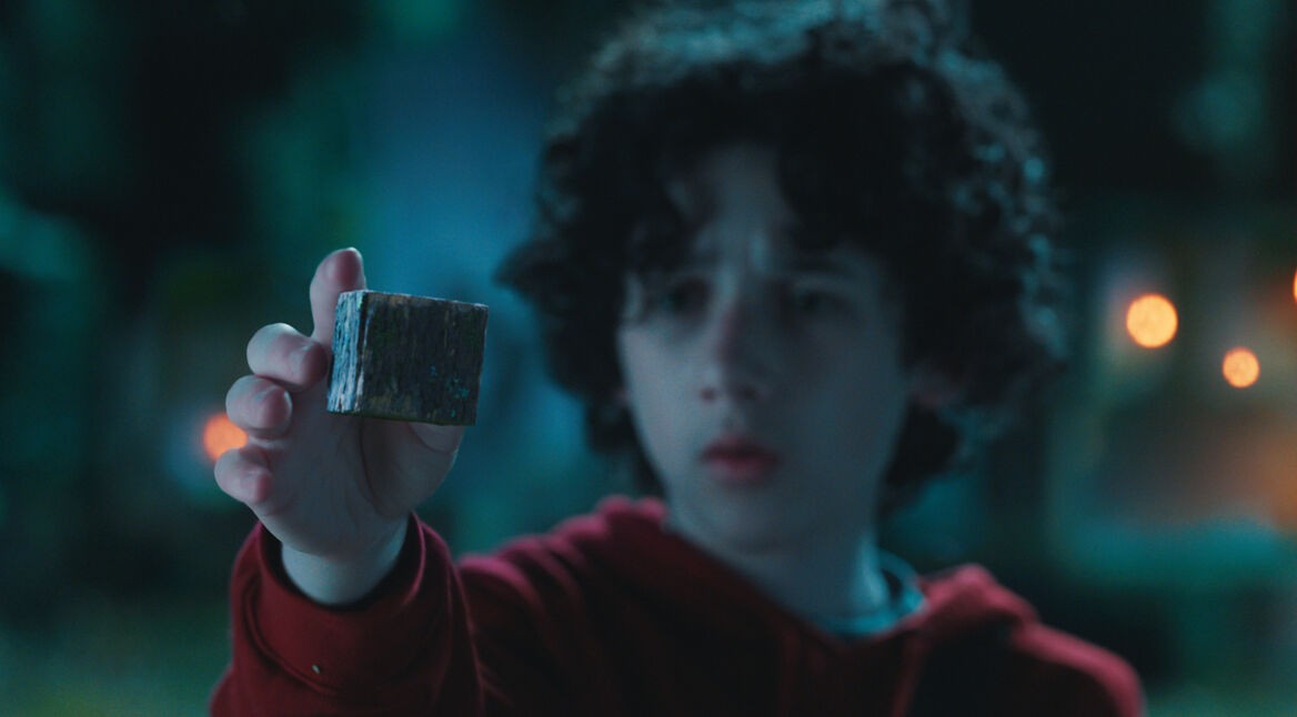 In dieser Szene aus Ein Minecraft Film ist Henry zu sehen, gespielt von Sebastian Hansen. Er trägt einen roten Hoodie und hält ein geheimnisvoll wirkendes, würfelförmiges Objekt aus Holz in die Kamera. Sein Blick ist konzentriert und leicht verwundert. Die mystische Lichtstimmung im Hintergrund verstärkt die geheimnisvolle Atmosphäre.