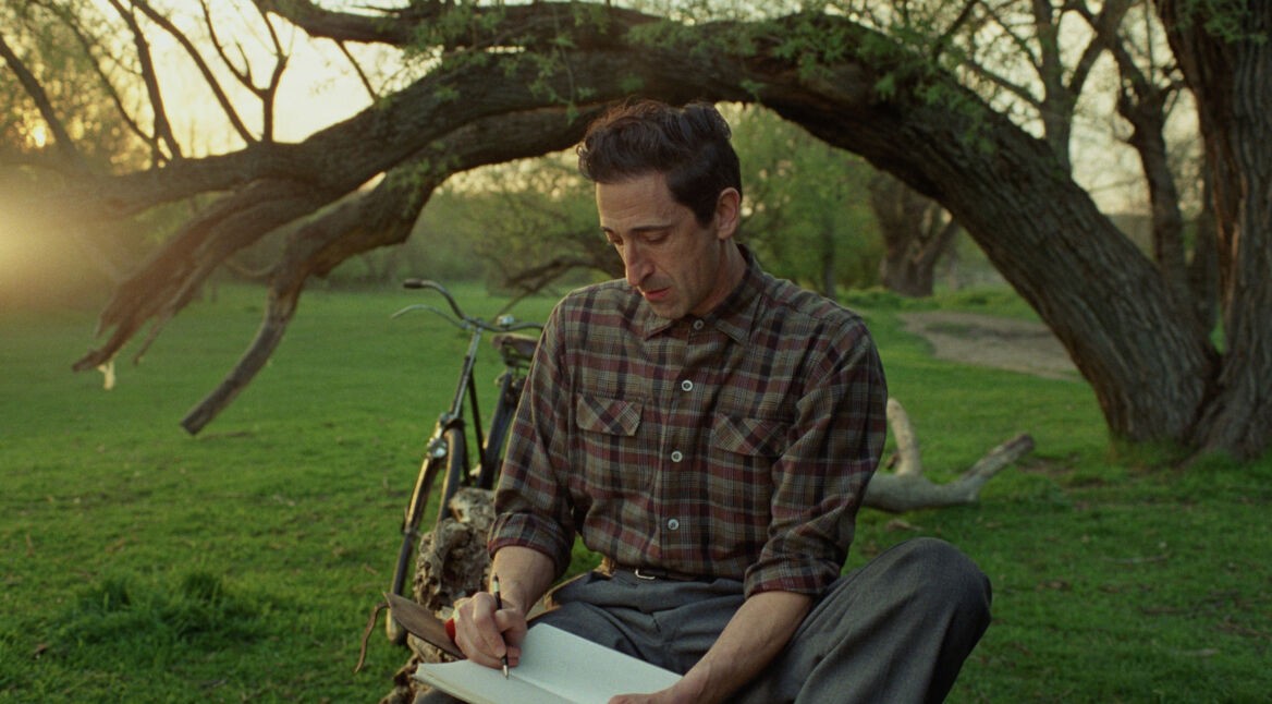 GigaTV-Highlights im April 2025: In einer Szene aus The Brutalist sitzt Adrien Brody als zurückhaltender Architekt in einem ruhigen Park auf dem Gras und schreibt in ein Notizbuch.