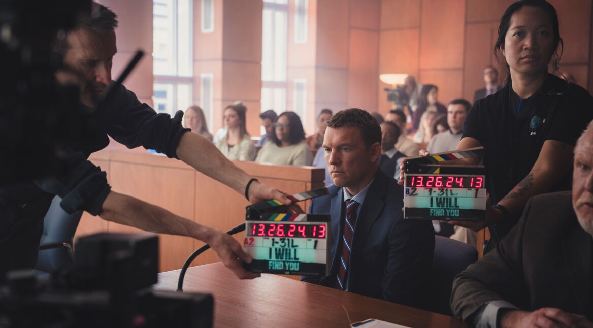 Auf diesem Bild zur Serie Nur für Dein Leben ist Sam Worthington als David Burroughs in einer Gerichtsszene zu sehen. Er sitzt ernst und fokussiert am Tisch, umgeben von Filmcrew-Mitgliedern mit Klappe, was auf eine laufende Produktion hinweist. Die Atmosphäre ist angespannt, mit Statisten im Hintergrund, die ein authentisches Gerichtssetting verstärken.