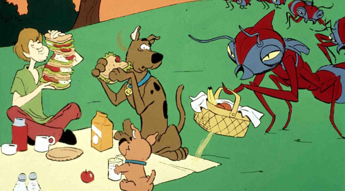 Auf dem Bild in dem Artikel zur Scooby-Doo-Live-Action-Serie ist eine Szene aus der klassischen Zeichentrickversion zu sehen: Shaggy sitzt im Gras und hält einen riesigen Sandwich-Turm, während Scooby-Doo und Scrappy-Doo auf einer Picknickdecke essen. Rechts greift eine riesige rote Ameise nach einem Picknickkorb. Die Stimmung ist skurril und leicht angespannt.