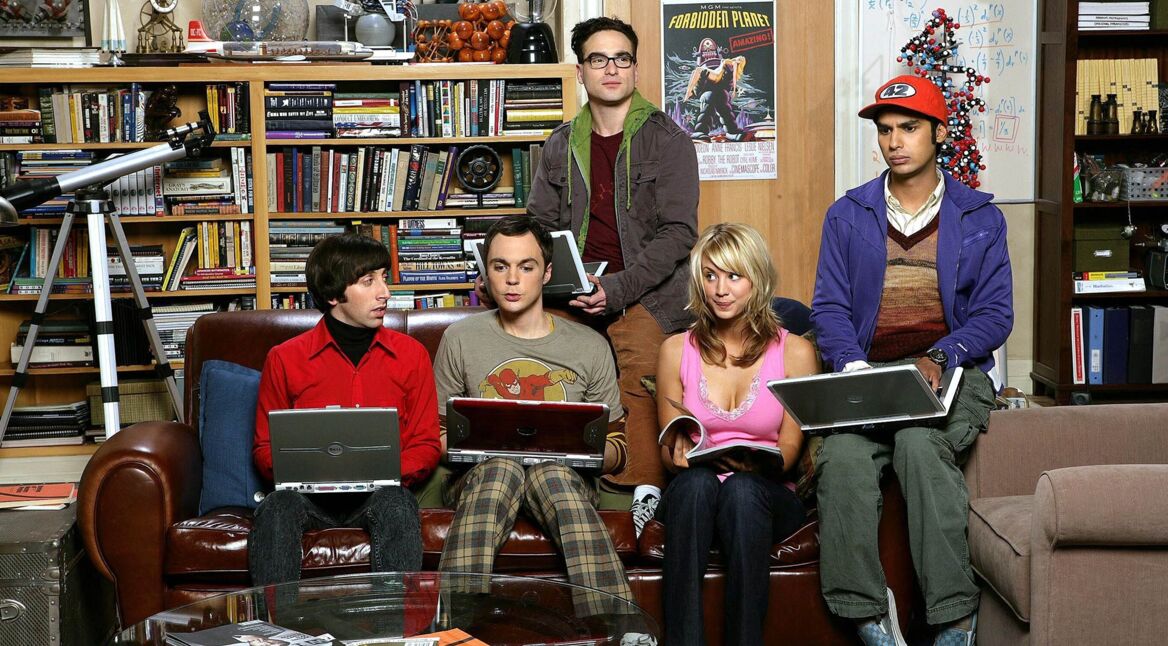Das Bild in dem Artikel zu den Serien wie The Big Bang Theory zeigt fünf Personen in einem Wohnzimmer mit Bücherregal und Teleskop. Sie sitzen eng beisammen auf einem Sofa und Stuhl, halten Laptops oder Bücher in den Händen. Die Szene wirkt freundlich und leicht nerdig, passend zum humorvollen und wissenschaftsaffinen Stil der Serie.