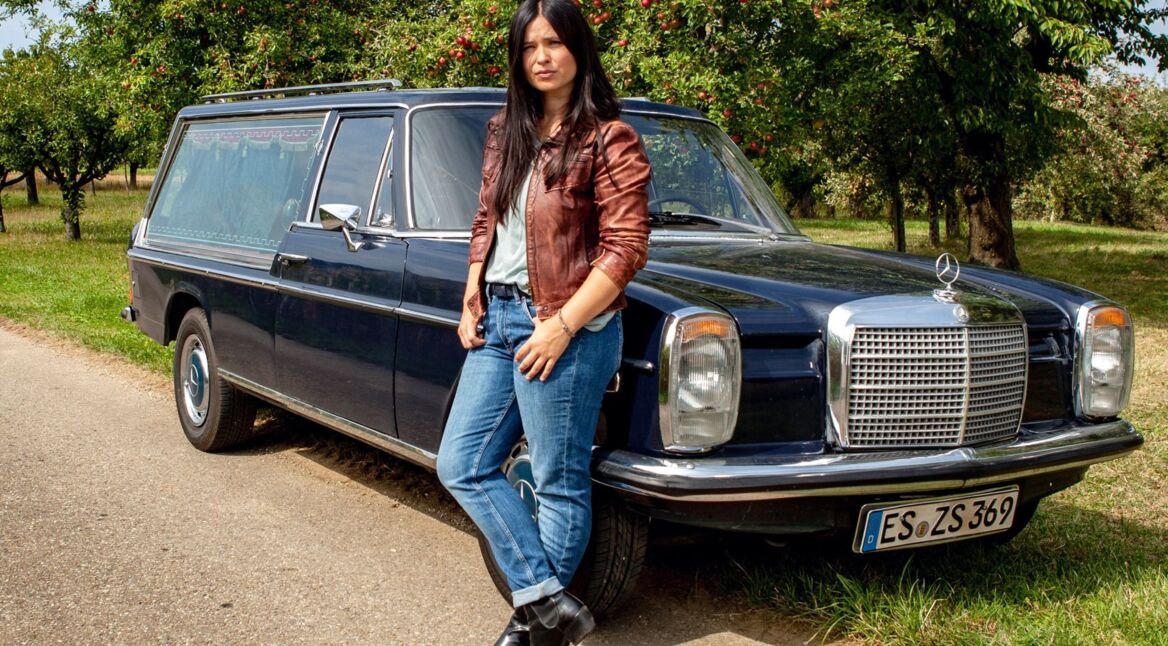 Anna Fischer lehnt in einer Szene von "Die Bestatterin" an einem Leichenwagen.