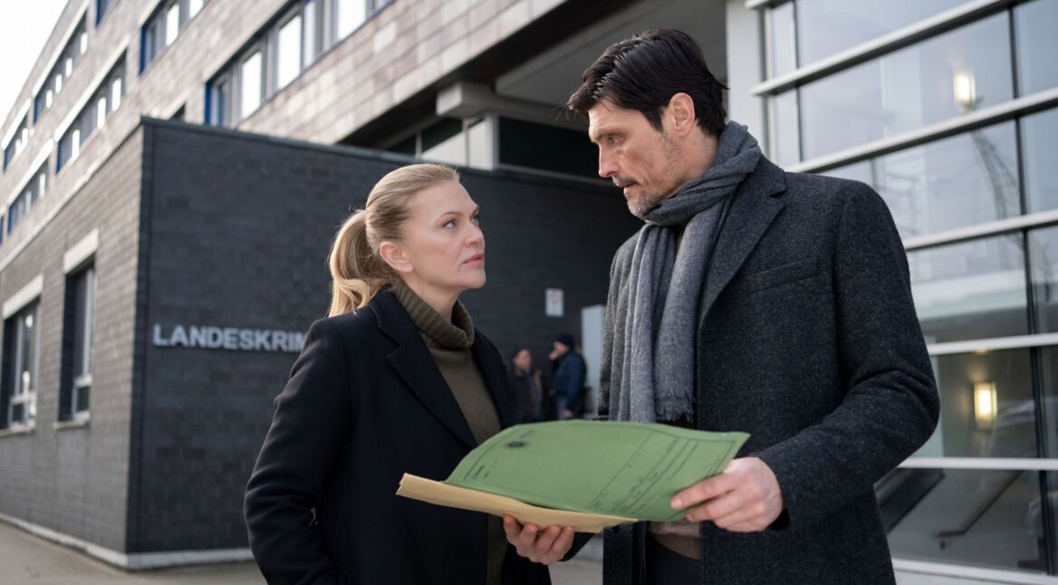 Die zwei Schauspieler Anna Loos und Stipe Erceg stehen vor dem Landeskriminalakt und schauen in eine Akte.