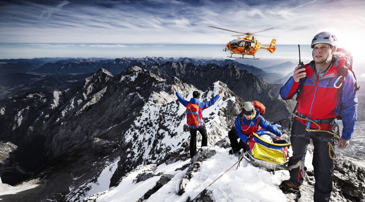 Drei Mitarbeiter:innen der Bergwacht, die sich mit einer geretteten Person auf einem Berggipfel befinden, weisen einen anfliegenden Hubschrauber ein.