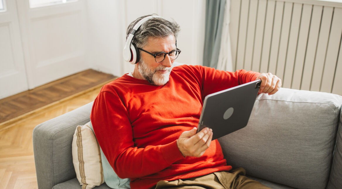 Ein älterer Mann mit grauem Haar und Bart sitzt entspannt auf einem hellgrauen Sofa und schaut mit Kopfhörern ein Video auf einem Tablet. Er trägt einen roten Pullover und wirkt konzentriert und zufrieden. Dieses Bild symbolisiert bequemes Streaming von zuhause und passt thematisch zur Darstellung der ARD Plus Kosten.