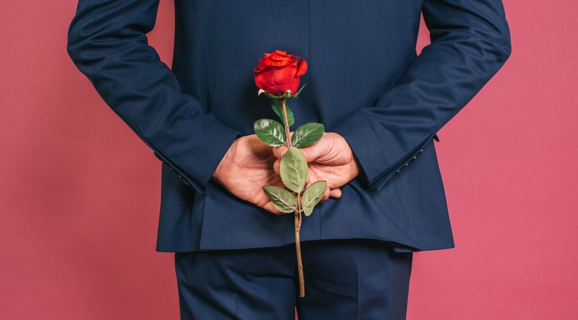 Auf dem Bild in dem Artikel zu den Bachelor-Formaten ist ein Mann in einem dunkelblauen Anzug von hinten zu sehen. Er hält eine rote Rose versteckt hinter seinem Rücken, während er vor einem pinkfarbenen Hintergrund steht.