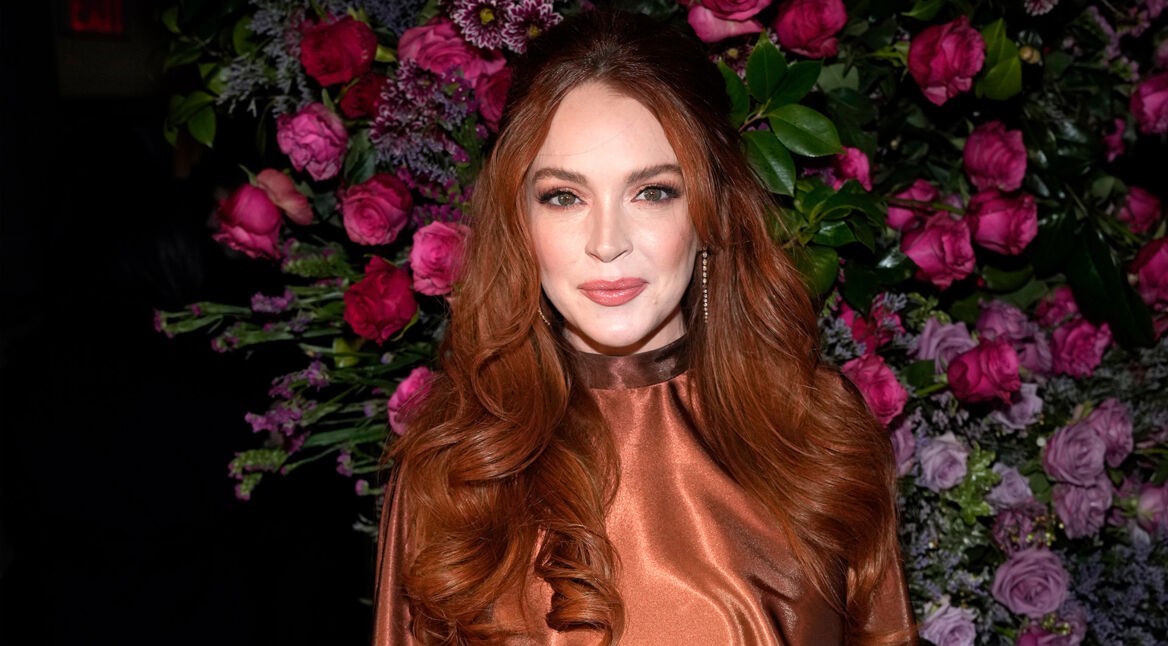 Auf dem Eventfoto ist Lindsay Lohan mit offenen, gewellten roten Haaren und einem schimmernden, bronzefarbenen Kleid zu sehen. Sie steht vor einer üppigen Blumenwand aus violetten und rosafarbenen Rosen und schaut direkt in die Kamera. Das Bild wird im Zusammenhang mit der möglicherweise geplanten Serie Count My Lies verwendet