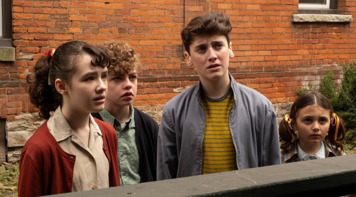 Welcome to Derry zeigt in dieser Szene vier Kinder, die mit ernsten und teils verunsicherten Blicken in dieselbe Richtung schauen. Die Gruppe steht vor einer Backsteinwand, ihre Kleidung wirkt retro und deutet auf eine frühere Zeitperiode hin.