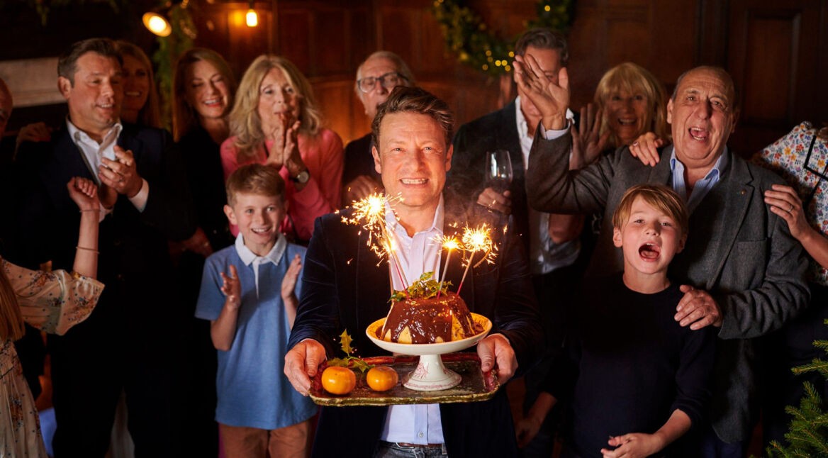 In dem Artikel zu den kostenlosen Jamie Oliver Folgen zu seinem 50. Geburtstag ist Jamie Oliver im Mittelpunkt einer festlichen Szene zu sehen. Er lächelt breit, hält einen Teller mit Schokopudding und Wunderkerzen in der Hand, während hinter ihm eine Gruppe aus Kindern und Erwachsenen applaudiert und feiert.