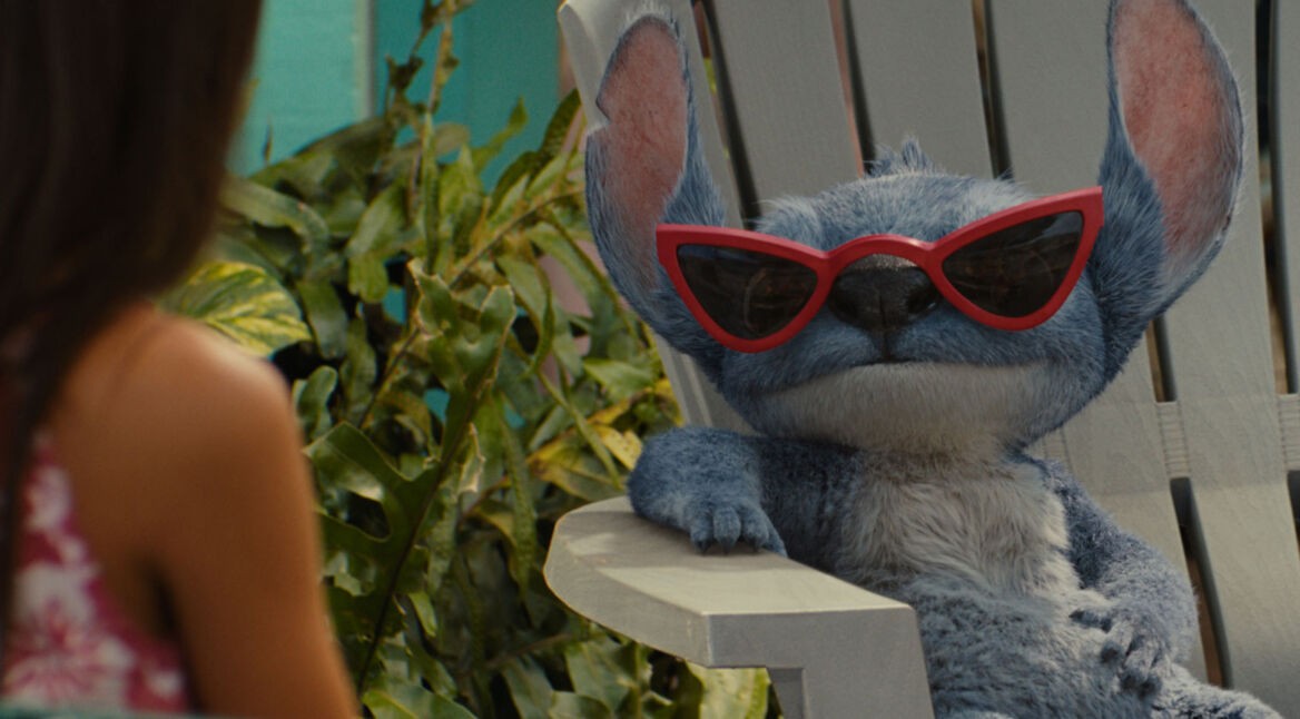 Auf dem Bild in dem Artikel zum Lilo & Stitch-Vergleich ist Stitch in der Live-Action-Version von 2025 in einer entspannten Szene zu sehen. Er sitzt lässig auf einem weißen Gartenstuhl, trägt eine rote Retro-Sonnenbrille und blickt cool zur Seite. Neben ihm ist unscharf Lilo zu erkennen.