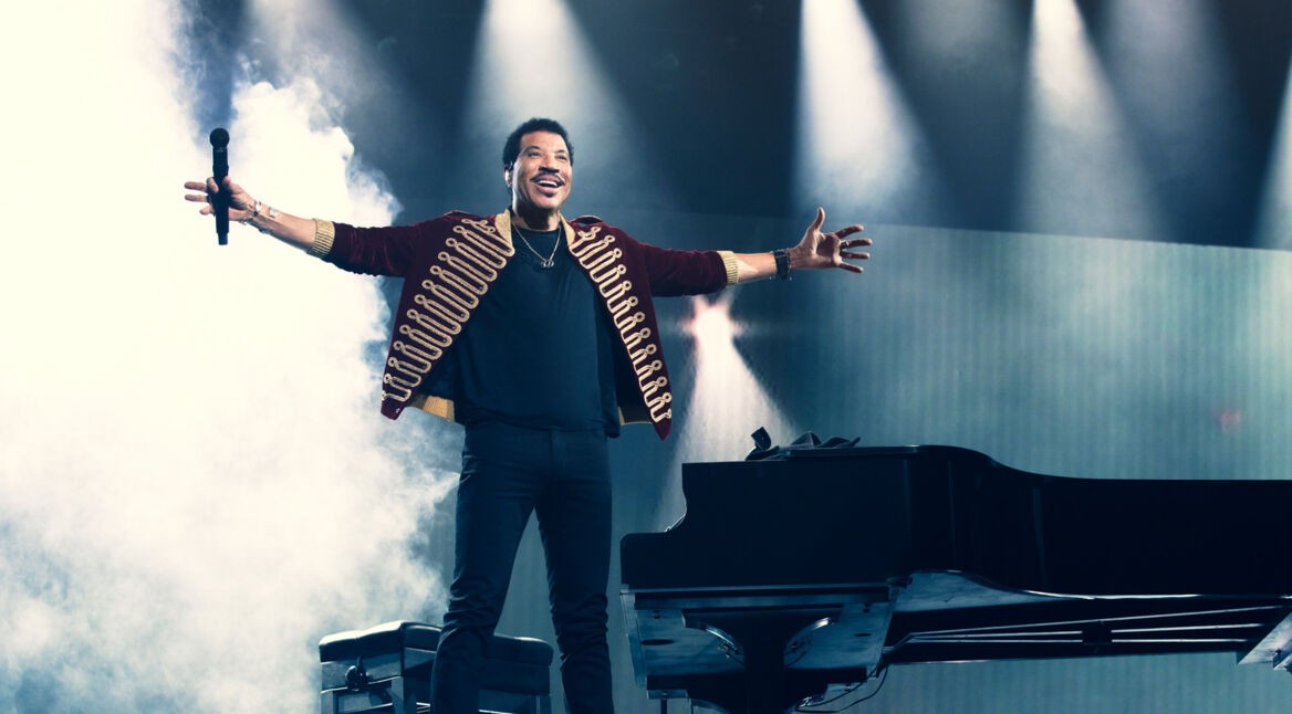 Im Artikel zu den Lionel Richie Tickets zeigt dieses Bild den Künstler in einem eindrucksvollen Moment auf der Bühne: Mit ausgebreiteten Armen steht Lionel Richie neben einem schwarzen Konzertflügel, umgeben von dichter Nebelstimmung und dramatischer Beleuchtung.