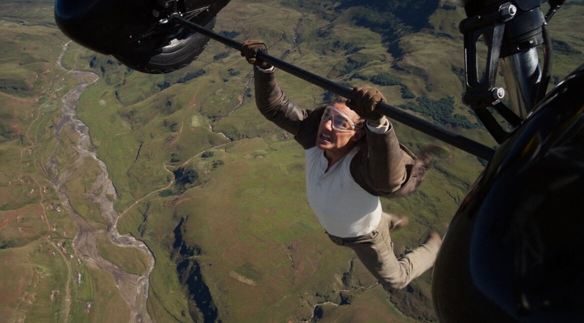 Im Bild zur Mission: Impossible 8 Kritik hängt Tom Cruise als Ethan Hunt in schwindelerregender Höhe an einem Helikopterfahrwerk über einer zerklüfteten Landschaft. Mit Schutzbrille und konzentriertem Gesichtsausdruck stemmt er sich gegen den Wind. Die Szene betont erneut die kompromisslose Action und den körperlichen Einsatz, den die Reihe berühmt gemacht hat.