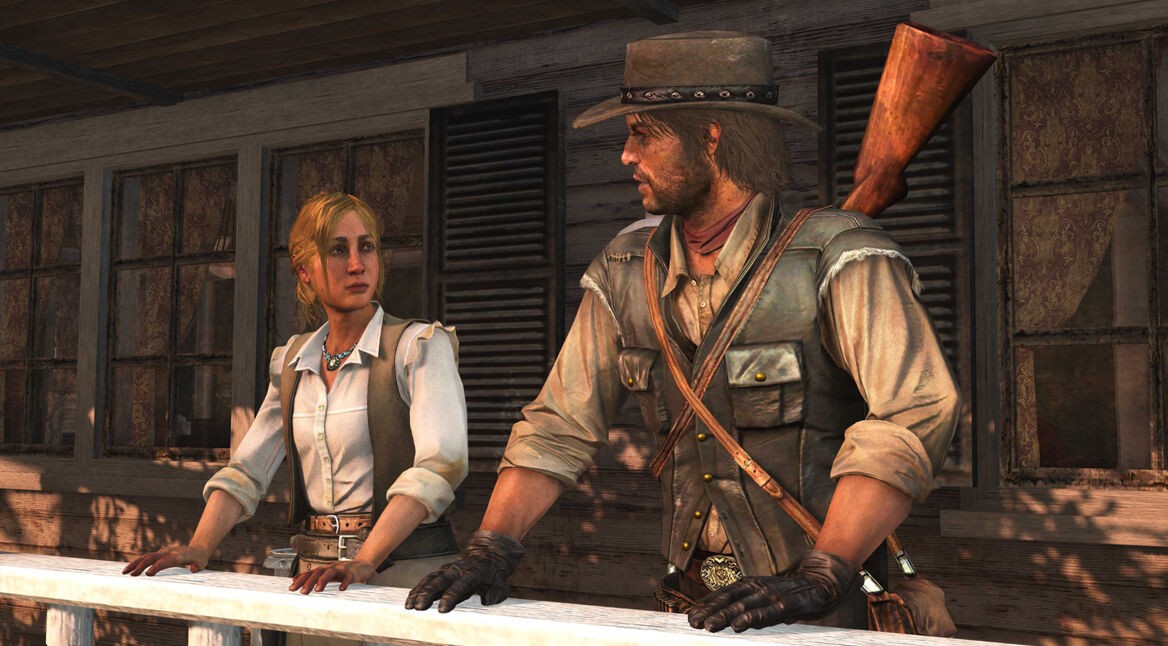 Im Screenshot aus dem Spiel Red Dead Redemption sind John Marston und seine Frau Abigail auf einer Holzveranda zu sehen. John trägt einen Cowboyhut, Handschuhe und eine Lederweste mit Gewehr auf dem Rücken, während Abigail in heller Bluse und Weste ernst schaut. Die Szene vermittelt eine ruhige, ernste Stimmung.