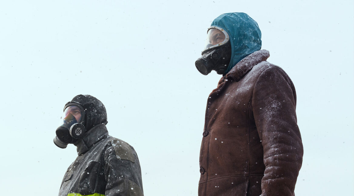 In dieser Szene aus Eternauta Staffel 1 stehen zwei Figuren in einer verschneiten Außenlandschaft mit Gasmaske und Winterkleidung. Links ist Favalli zu sehen, gespielt von César Troncoso, in dunkler Jacke mit reflektierendem Streifen. Rechts steht Juan, dargestellt von Ricardo Darín, in brauner Felljacke und Kapuze. Beide blicken ernst in die Ferne – die Stimmung ist angespannt und bedrohlich.