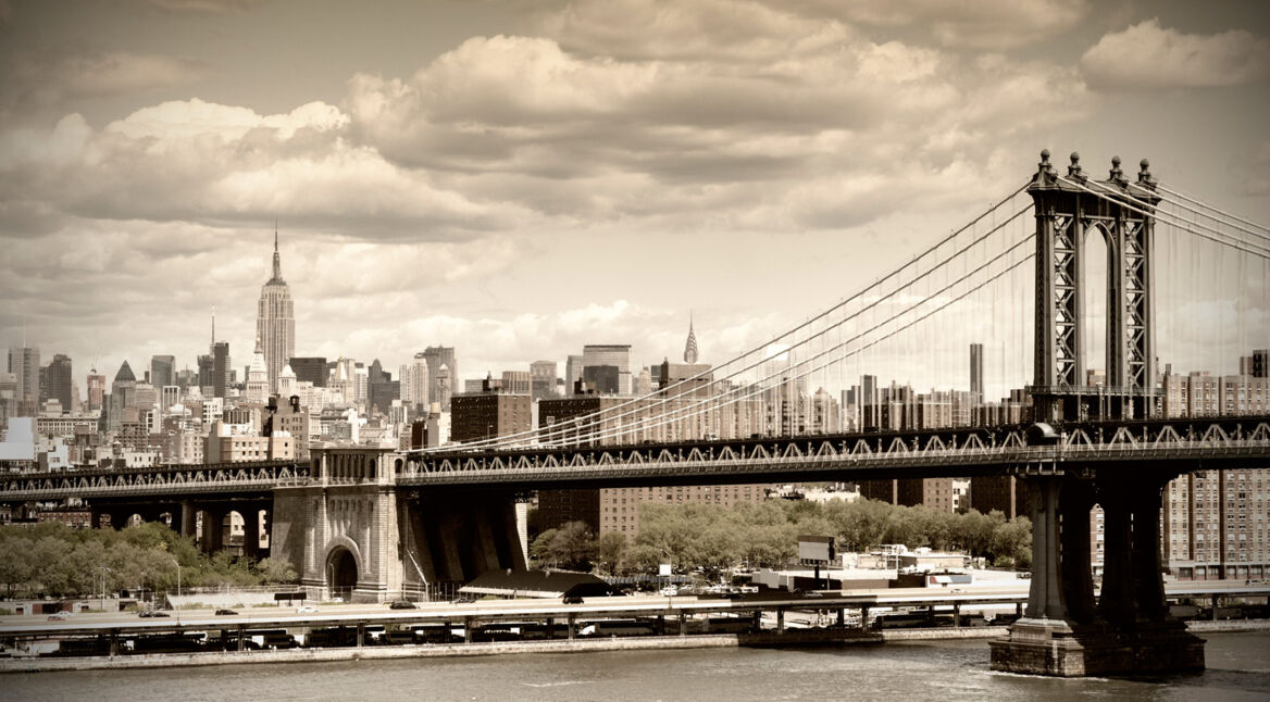 Auf diesem Symbolbild zur Zeit der Unschuld-Serie ist die Manhattan Bridge im Vordergrund zu sehen, während im Hintergrund die Skyline von New York mit dem Empire State Building zu sehen ist. Das Bild ist in sepiafarbenem Ton gehalten und vermittelt eine nostalgische, historische Atmosphäre, die an das New York der 1920er Jahre erinnert.