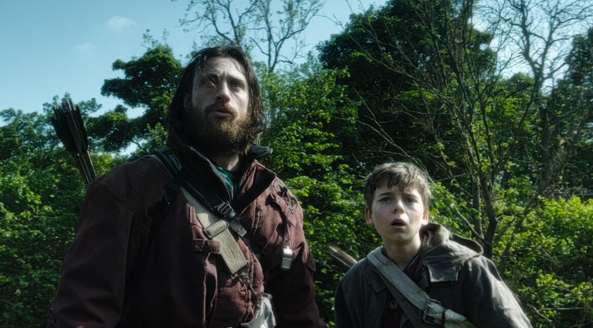 In dieser Szene aus 28 Years Later stehen Jamie, gespielt von Aaron Taylor-Johnson, und sein Sohn Spike, gespielt von Alfie Williams, inmitten eines dichten Waldes. Beide blicken alarmiert nach oben, mit ernsten, angespannten Gesichtern. Jamie trägt einen Köcher mit Pfeilen auf dem Rücken