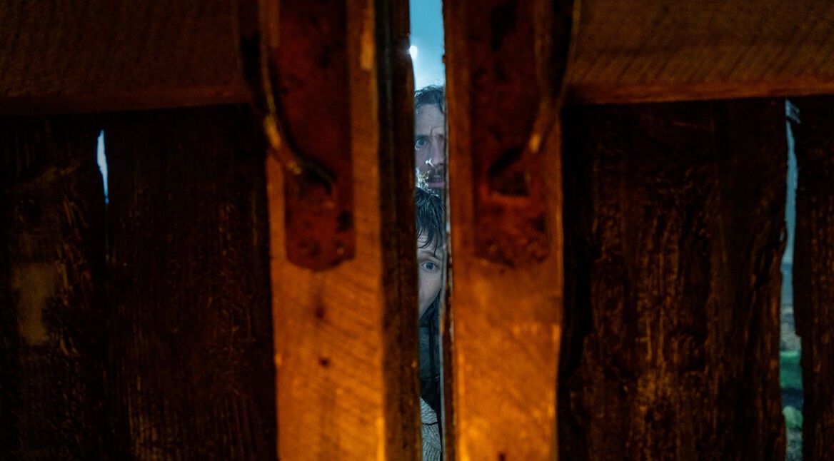 Das Bild aus der Filmkritik zu 28 Years Later zeigt eine intensive Szene mit Jamie (Aaron Taylor-Johnson) und Spike (Alfie Williams), die durch einen schmalen Spalt in einer massiven Holztür blicken. Ihre Gesichter wirken angespannt und vorsichtig, während nur wenig Licht ins Bild fällt – die bedrohliche Stimmung unterstreicht das post-apokalyptische Setting eindrucksvoll.