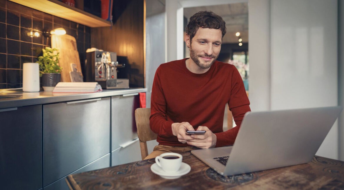 Vodafone E-Mail & Cloud im Netzwelt-Test: Mann in roten Pullover sitzt mit einem Kaffee vor seinem Laptop in einer modern eingerichteten Küche.