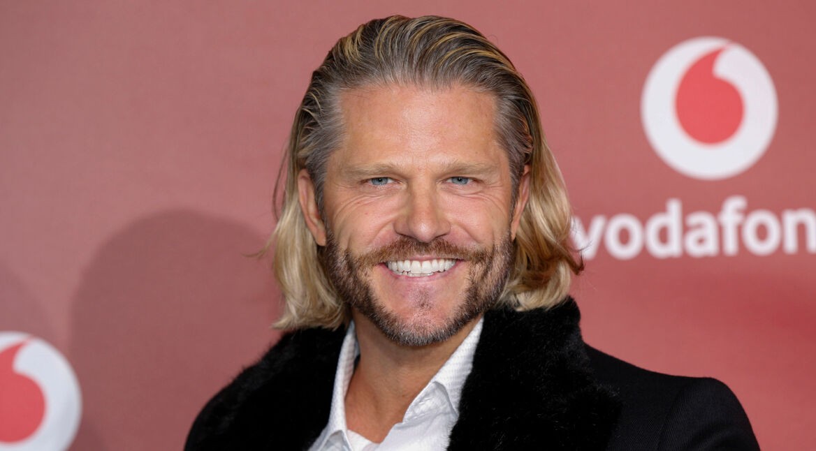 Auf dem Bild zur Übersicht Alle Bachelor ist Paul Janke vor einer roten Fotowand mit Vodafone-Logo zu sehen. Er trägt ein schwarzes Jackett mit Fellkragen und ein weißes Hemd. Mit einem strahlenden Lächeln und zurückgekämmten blonden Haaren wirkt er selbstbewusst und charmant – ganz im Stil des wohl bekanntesten Junggesellen der Sendung.