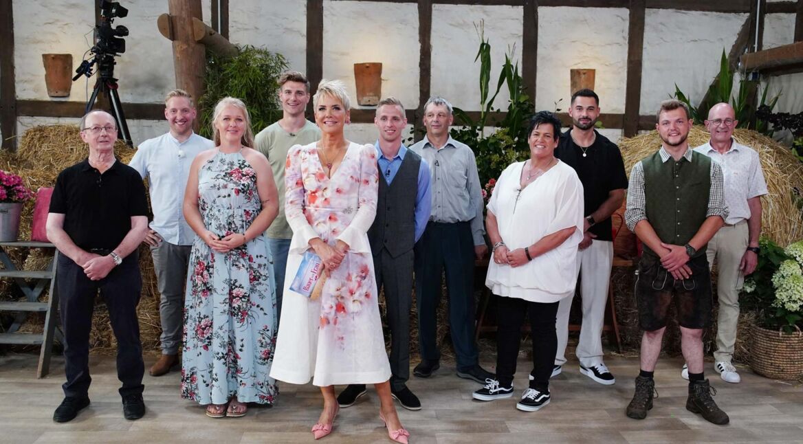 Auf dem Bild zur Sendung Bauer sucht Frau 2025 stehen die Moderatorin Inka Bause und die Teilnehmer:innen Walter, Daniel, Insa, Friedrich, Christopher, Johann, Simone, Basti, Michi und Herbert in einem rustikal dekorierten Studio mit Strohballen und Blumen.