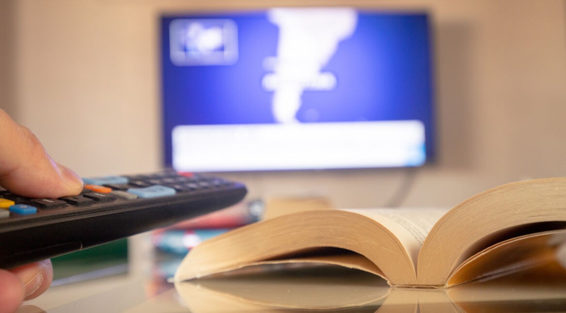 Ein aufgeschlagenes Buch liegt im Vordergrund auf einem Glastisch, während im Hintergrund unscharf ein Fernseher mit Nachrichtenbildschirm zu sehen ist. Eine Hand hält eine Fernbedienung in Richtung TV. Das Bild symbolisiert die Verbindung von Literatur und Fernsehen und steht stellvertretend für die kommenden Buchverfilmungen 2025, bei denen Romane zum Leben erweckt werden.