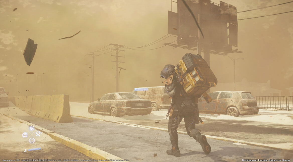 Auf diesem Screenshot aus Death Stranding 2: On the Beach kämpft der Lieferant in schwerer Ausrüstung gegen einen heftigen Sandsturm an. Er geht gebückt und schützt sein Gesicht, während hinter ihm verlassene, verrostete Autos und eine staubige Straße eine postapokalyptische Stimmung erzeugen.