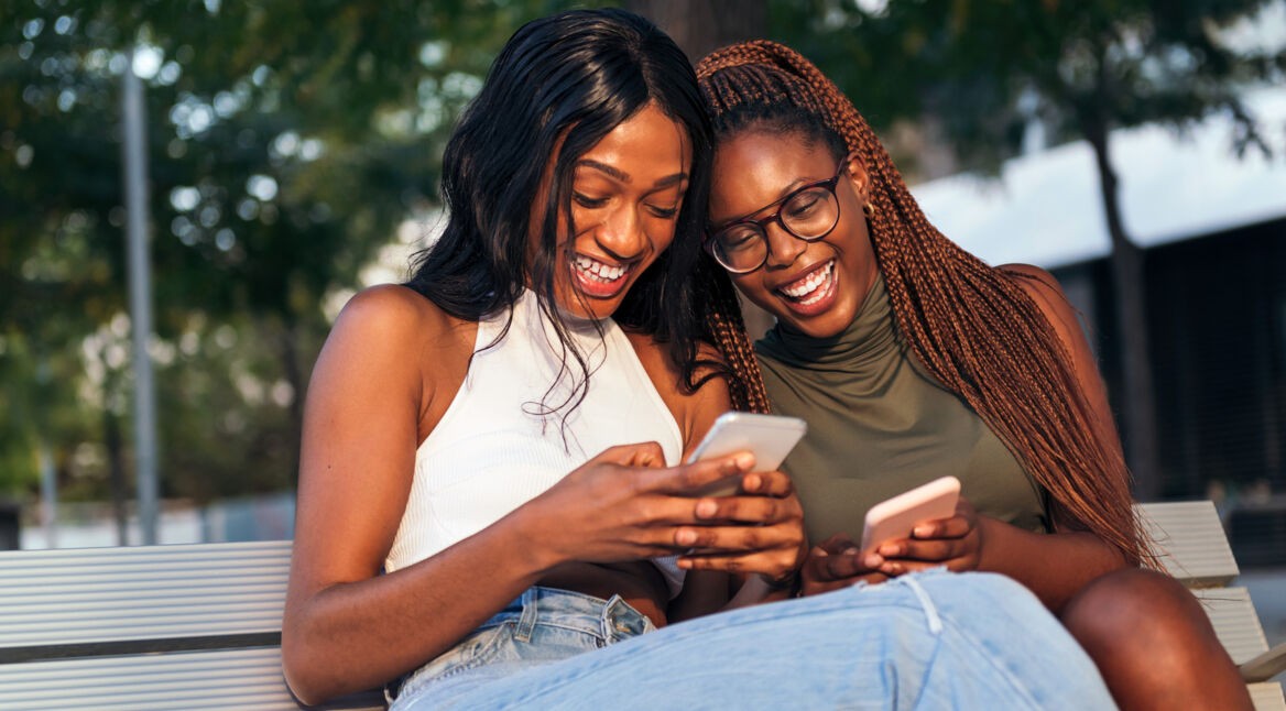 Zwei junge Frauen sitzen auf einer Bank, schauen auf ihre Smartphones und lachen.