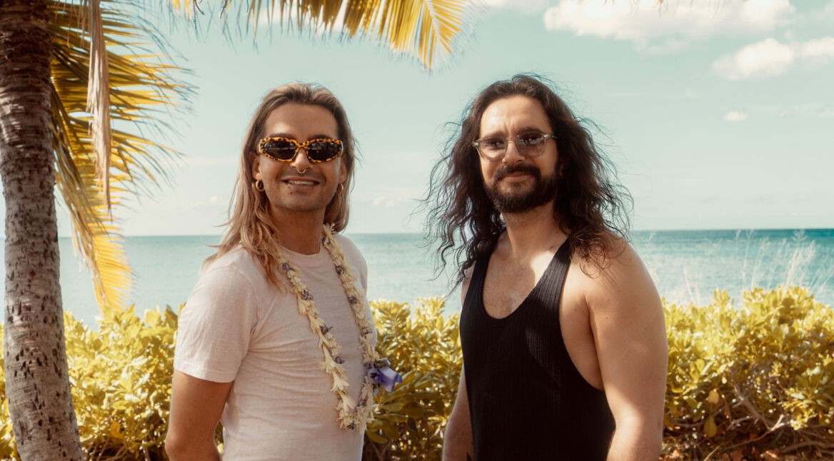 Auf dem Bild zur möglichen Fortsetzung Kaulitz & Kaulitz Staffel 3 stehen Bill und Tom Kaulitz entspannt vor tropischer Kulisse mit Meerblick. Bill trägt eine auffällige Sonnenbrille, ein weißes Shirt und eine Blumenkette, während Tom mit langem Haar und schwarzem Tanktop lässig wirkt. Die Stimmung ist sommerlich und gelöst – ein symbolischer Ausblick auf neue Folgen.