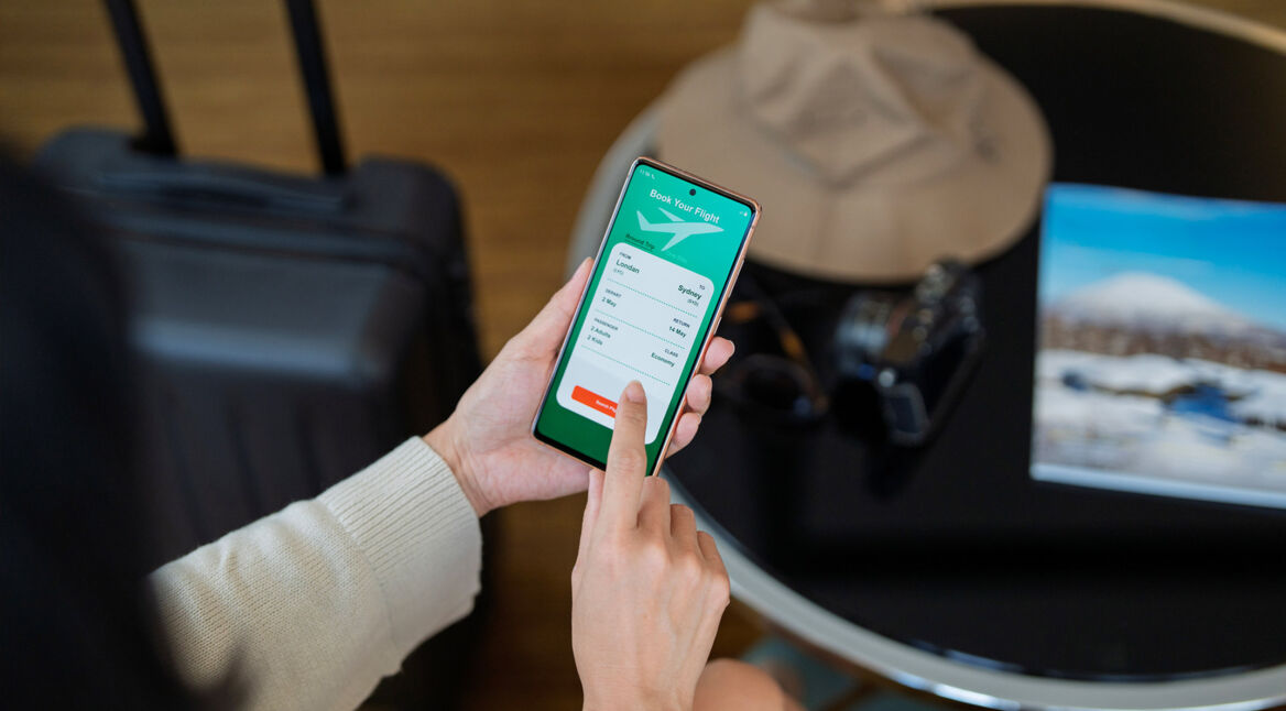 Eine Frau hält ein Smartphone in der Hand und nutzt eine der Reiseplanungs-Apps, um einen Flug von London nach Sydney zu buchen. Im Hintergrund stehen ein schwarzer Koffer, ein Sonnenhut und eine Kamera auf einem runden Tisch, daneben liegt ein Reisemagazin. Die Szene vermittelt Vorfreude und Reiselust.