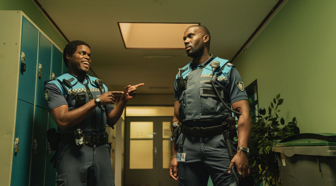 Bild aus Almost Cops: Zwei Hilfspolizisten in Uniform stehen sich in einer Umkleide gegenüber. Während der eine grinsend auf den anderen zeigt, blickt ihn dieser wütend an.