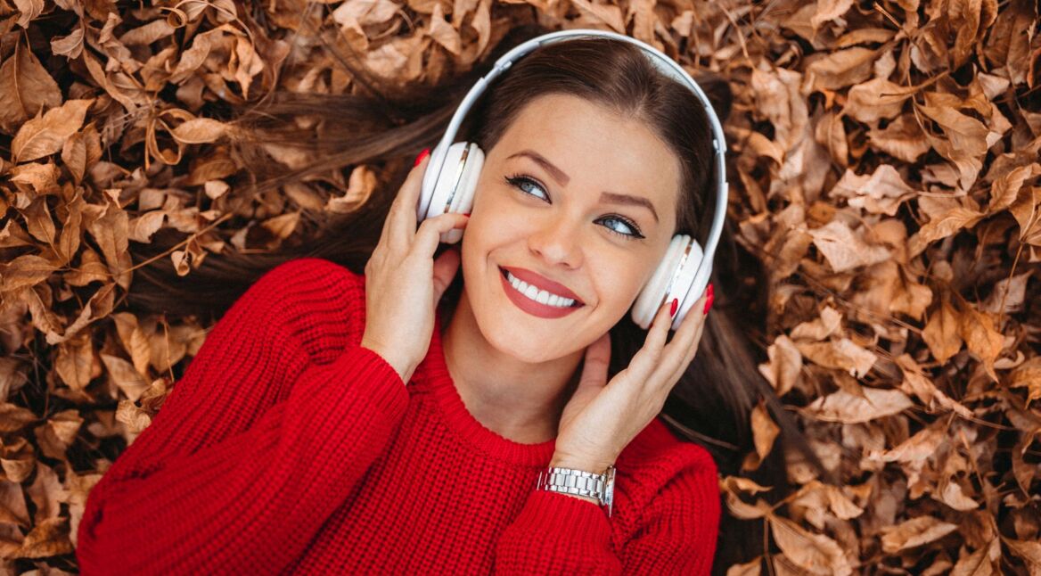 Eine Frau mit Over-Ear-Kopfhörern liegt auf einem Boden inmitten von herabgefallender Herbstblätter.