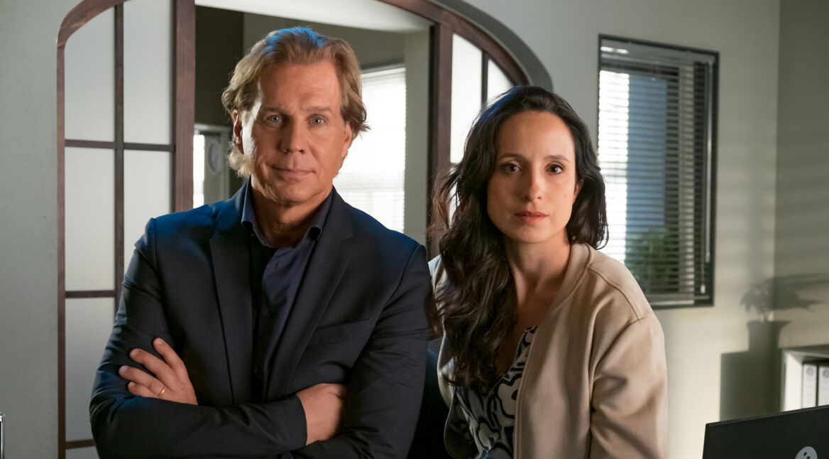 Auf dem Bild zu den Der Alte-Darsteller:innen sind Thomas Heinze als Hauptkommissar Caspar Bergmann (rechts) und Stephanie Stumph als Kommissarin Annabell Lorenz (links) in einem Büro zu sehen. Heinze steht mit verschränkten Armen, trägt ein dunkles Sakko und schaut ruhig in die Kamera, während Stumph in heller Jacke ernst und aufmerksam daneben sitzt.