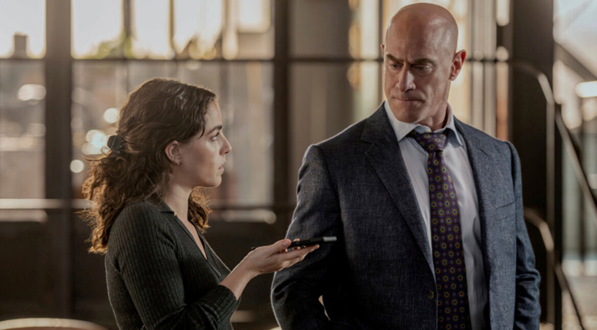 In den GigaTV-Highlights im August ist eine Szene aus Law and Order: Organized Crime zu sehen, in der Elliot Stabler, gespielt von Christopher Meloni, im Anzug einer jungen Frau mit Aufnahmegerät gegenübersteht.