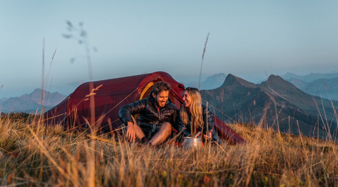 Auf diesem Bild der GO Real Life-Kampagne sitzen ein Mann und eine Frau entspannt im Gras vor einem roten Nordisk-Zelt. Die beiden lachen sich an und genießen die Aussicht auf eine Berglandschaft bei Sonnenaufgang oder Sonnenuntergang. Die Szene vermittelt Ruhe, Naturverbundenheit und echte Outdoor-Erlebnisse.