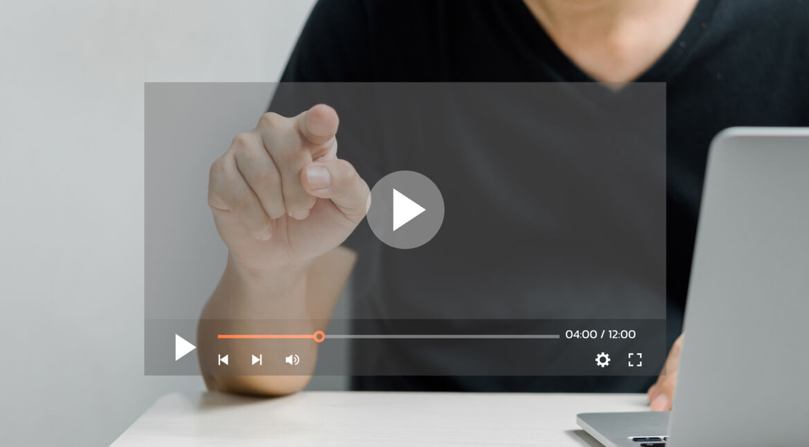 Ein Mann in schwarzem T-Shirt sitzt an einem Tisch und zeigt mit dem Finger auf einen transparent eingeblendeten Play-Button eines Videoplayers. Auf der rechten Seite ist ein aufgeklappter Laptop zu sehen. Das Bild symbolisiert die Nutzung eines digitalen Tools und steht stellvertretend für das Thema Sora Videos erstellen.