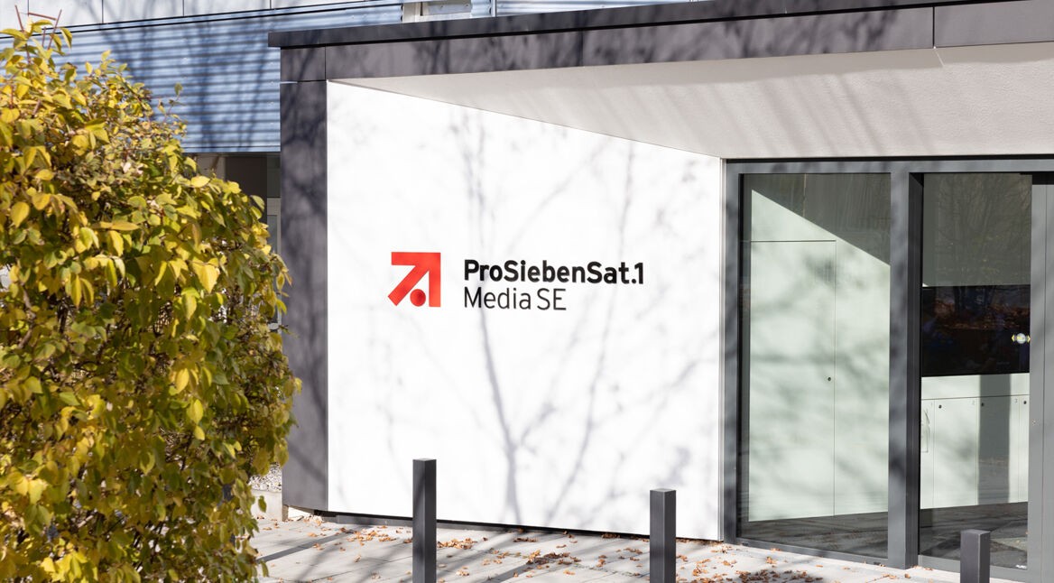 Auf dem Bild zur angekündigten Reality-Show The Hunt ProSieben ist der Eingangsbereich des ProSiebenSat.1 Media SE-Gebäudes bei Tageslicht zu sehen. Das moderne Gebäude zeigt das bekannte rot-schwarze Senderlogo auf weißem Hintergrund. Im Vordergrund befindet sich ein herbstlicher Baum mit gelbem Laub.