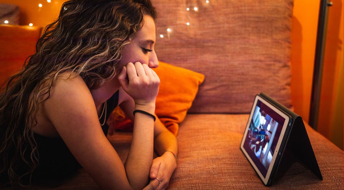 Ein junges Mädchen mit langen, gewellten Haaren liegt entspannt auf einem orangefarbenen Sofa und schaut konzentriert ein YouTube-Video auf einem Tablet. Die warme Beleuchtung mit kleinen Lichterketten im Hintergrund sorgt für eine gemütliche Stimmung. Die Szene steht exemplarisch dafür, ein Video ohne die automatischen YouTube-Übersetzungen anzuschauen.