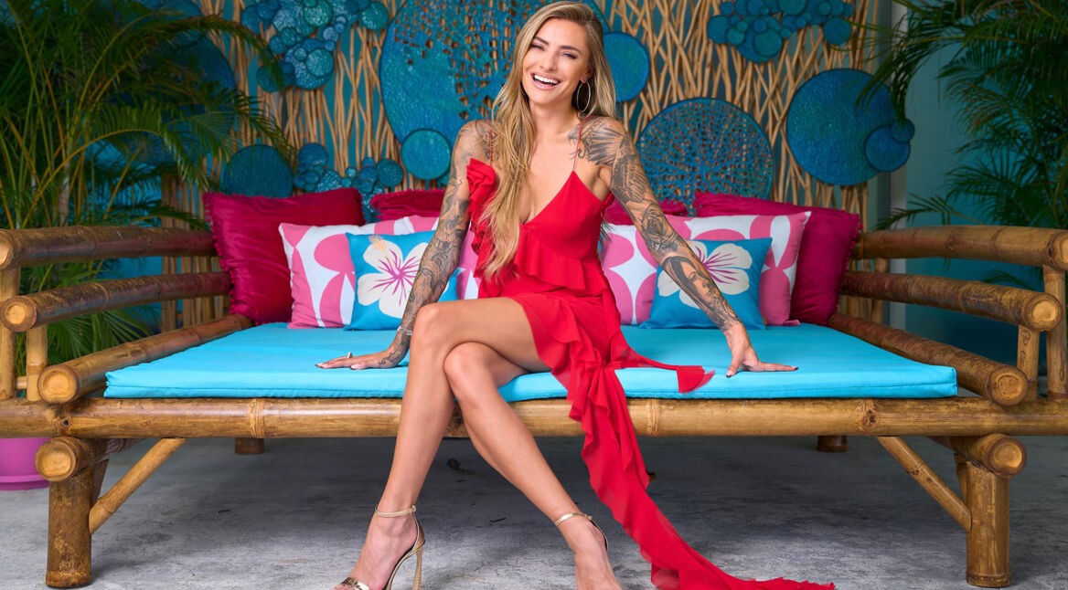 Moderatorin Sophia Thomalla sitzt lachend auf einer türkisfarbenen Couch. Sie trägt ein rotes Kleid mit goldene High Heels. Das Bild ist ein Promo-Bild für Are You The One Realitystars in Love Staffel 5.