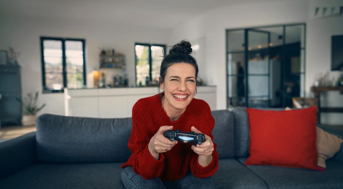 PS5 Upload Probleme: Frau sitzt freudig auf einem Sofa und hat einen PlayStation-Controller in der Hand.