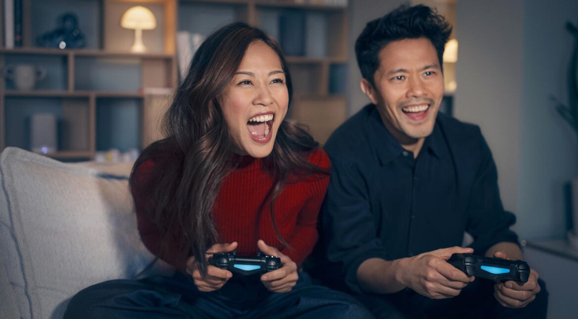 PS5 WLAN Probleme: Ein Mann und eine Frau sitzen aufgeregt auf einem Sofa und halten PlayStation Controller in der Hand.